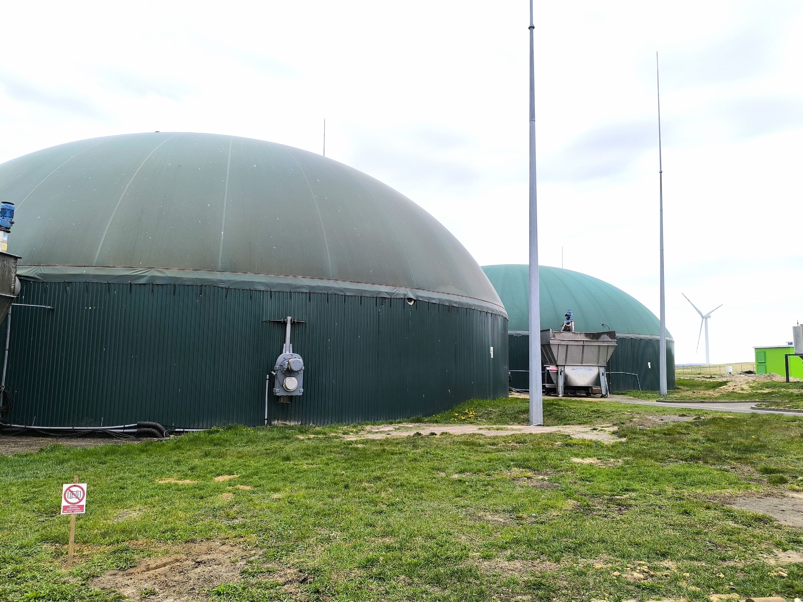 Zbiorniki instalacji biogazowej z zielonymi kopułami, z turbiną wiatrową w tle.